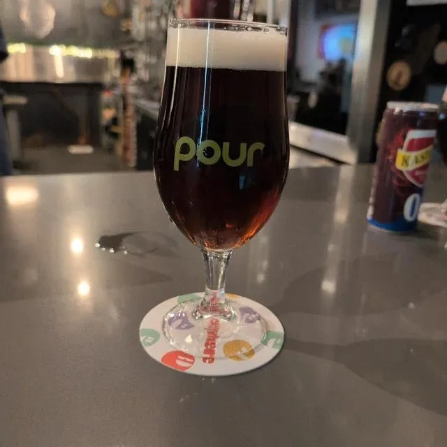 Stemmed glass of frothy beer with 'pour' text on colorful coaster.