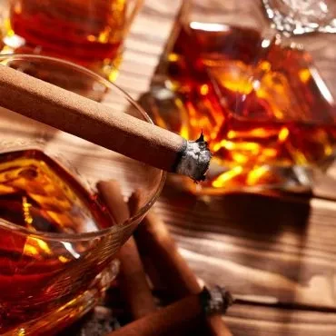 Warm photo of lit cigar on glass of amber liquid, cigars nearby.