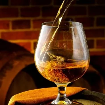 Golden-amber spirit pouring into a snifter glass, rustic barrels.