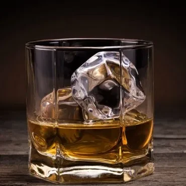 Amber whiskey in faceted glass with large ice on rustic wood.