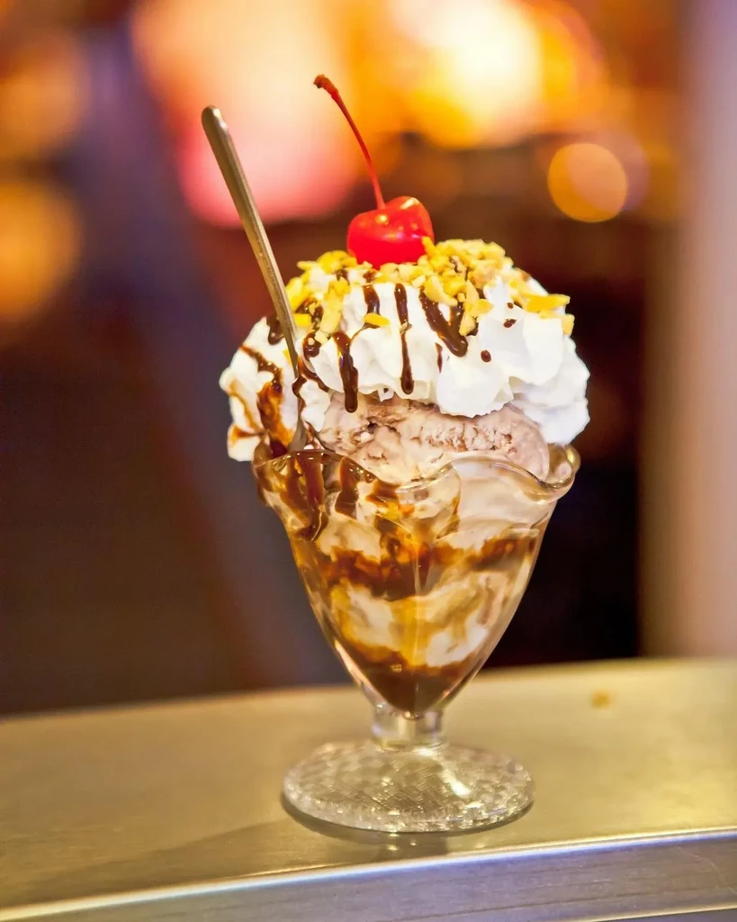 Ice cream sundae in glass, topped with cream, chocolate, nuts, cherry.