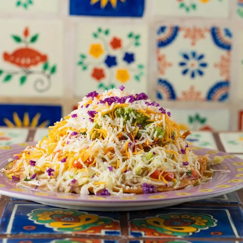 Piled high colorful dish with cheese, guacamole, lettuce, on patterned tiles.