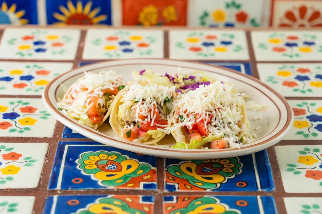 Three tacos with cheese, tomatoes, lettuce on a colorful tiled table.