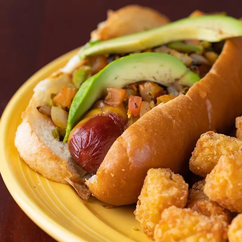 Gourmet hot dog with avocado and diced vegetables, served with tater tots.