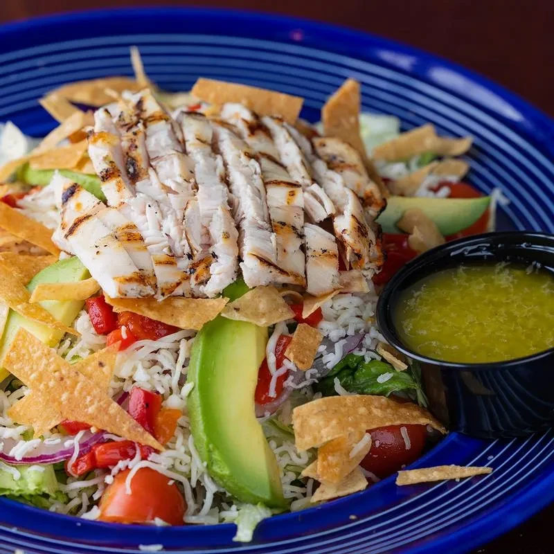 Grilled chicken salad, avocado, and dressing in a blue bowl.