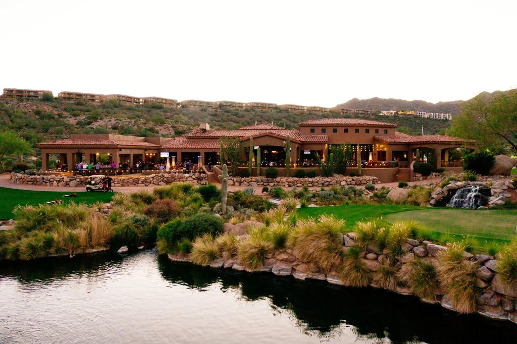 Spanish-colonial clubhouse patio with people, golf course, and desert pond.