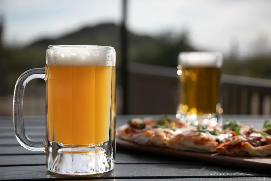 Clear beer mug with frothy head, blurred pizza in outdoor setting.