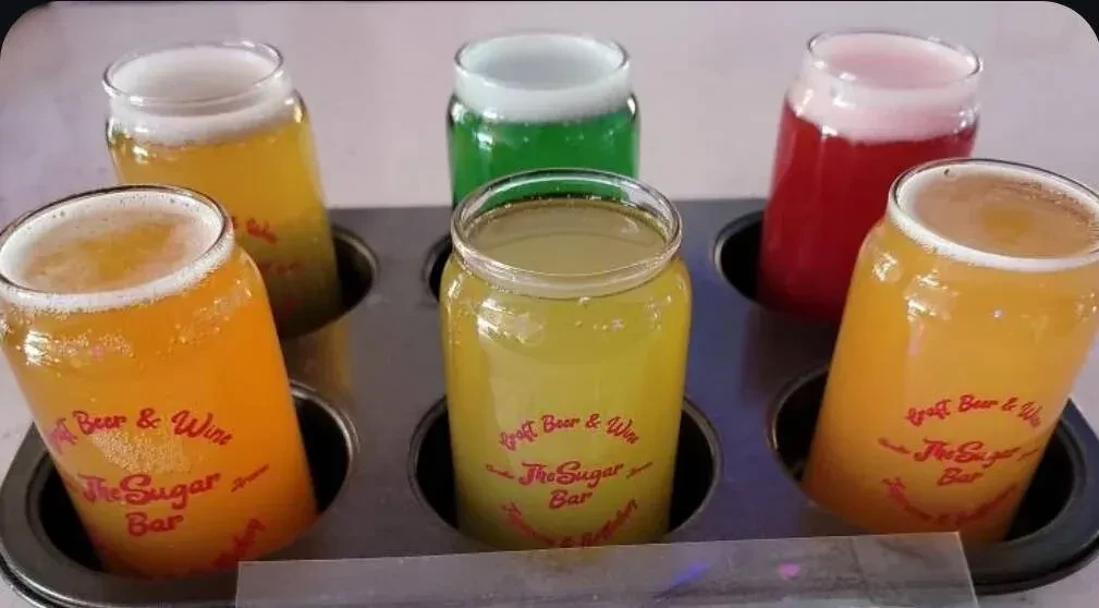 A flight of six can-shaped glasses filled with colorful, frothy drinks.
