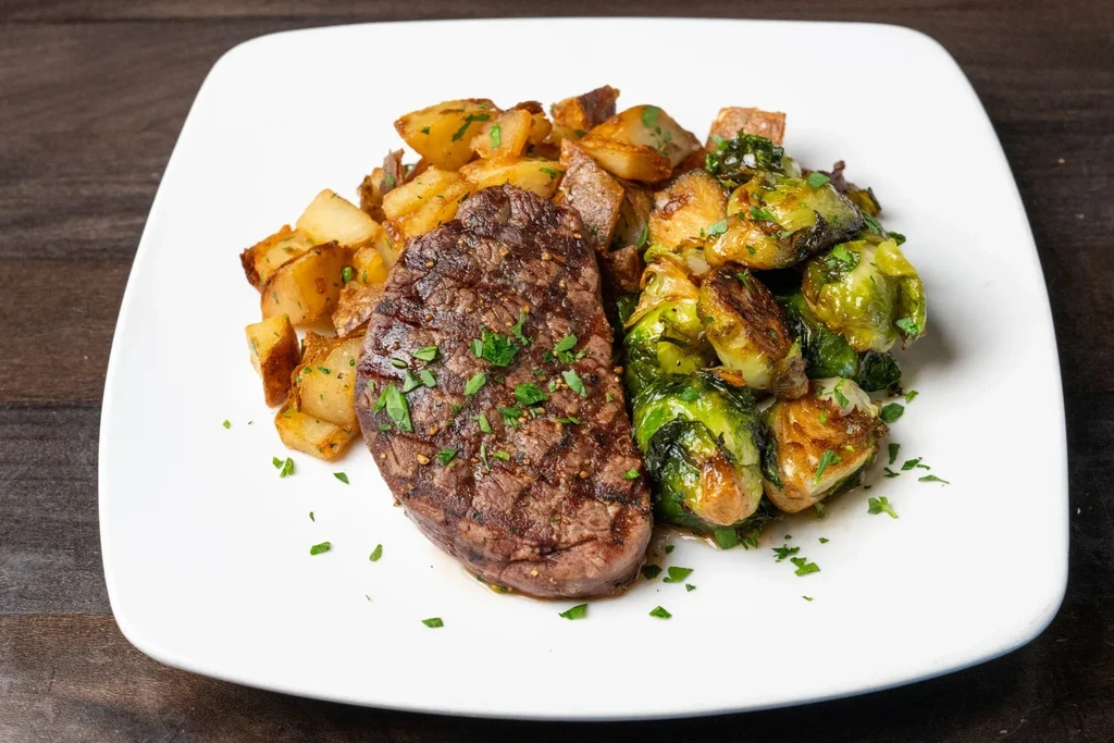 Medium-rare steak, roasted potatoes, Brussels sprouts, garnished on white plate.