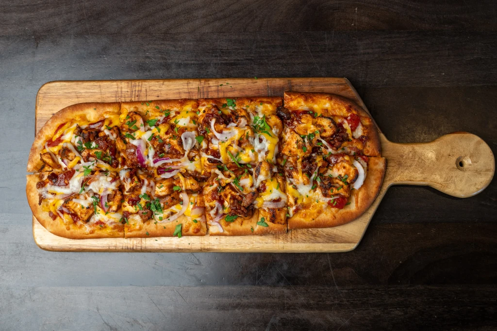 Pre-cut chicken flatbread pizza with cheese and herbs on a wooden board.