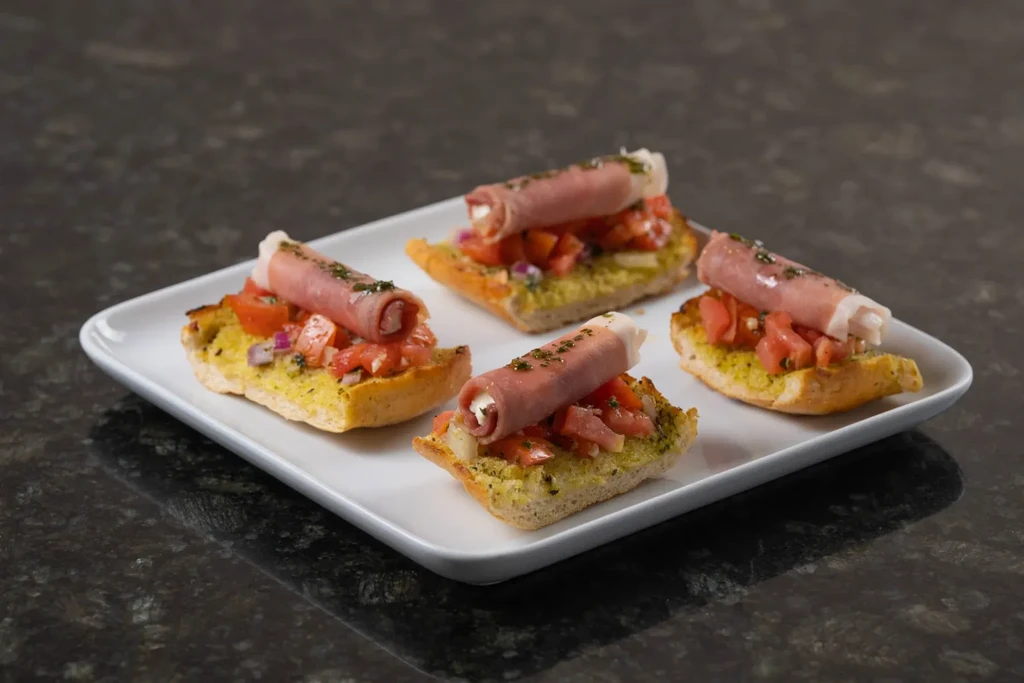 Four bruschetta with pesto, tomatoes, onions, and filled prosciutto rolls.