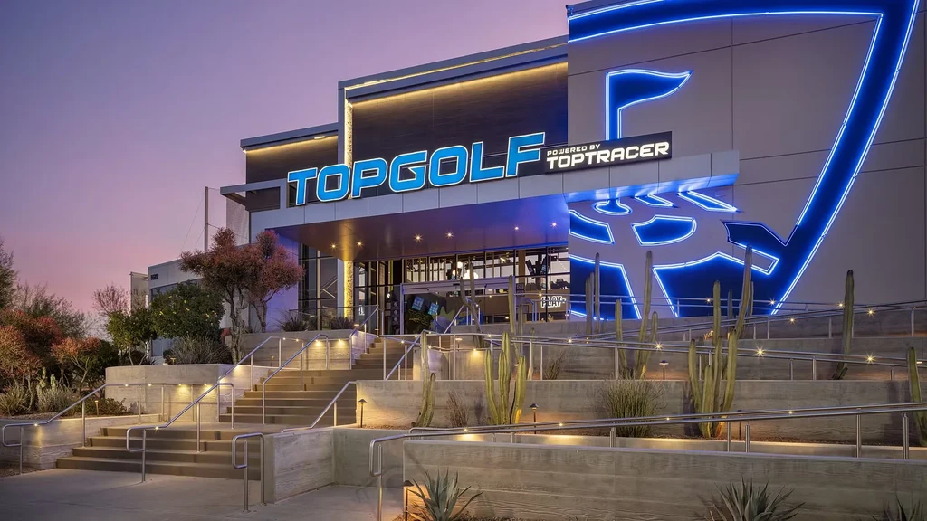 Modern Topgolf complex exterior at dusk with blue neon sign and desert landscaping.