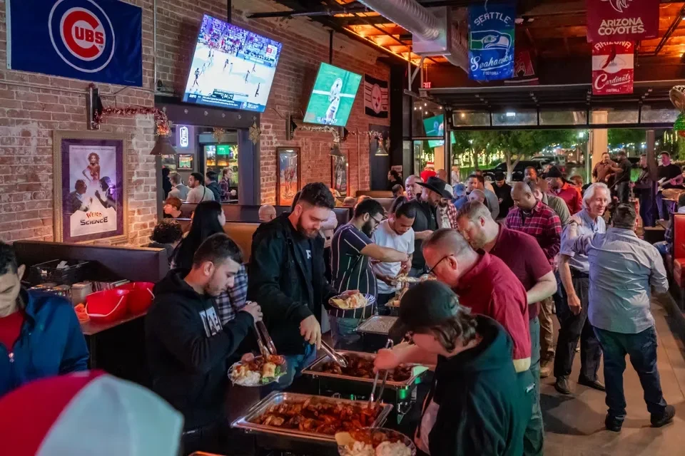Busy sports bar buffet with people, sports flags, and multiple TVs.