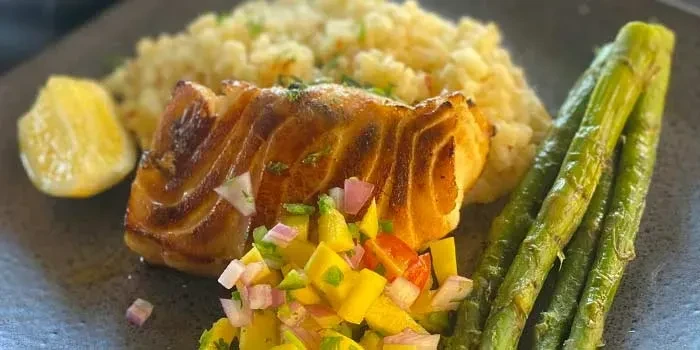 Plated golden-brown fish with mango salsa, rice, asparagus, and lemon.