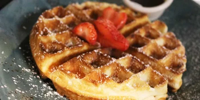 Golden-brown waffle with strawberries and powdered sugar on a dark plate.