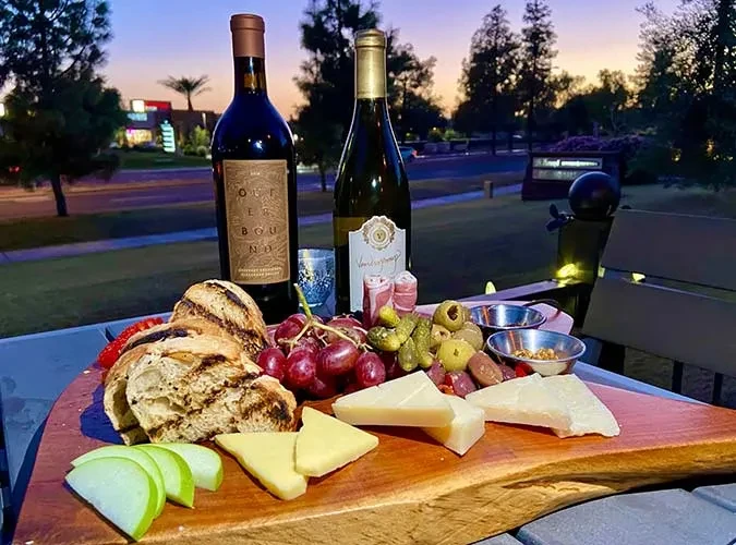 Charcuterie board with cheese, grapes, and wine bottles outdoors at dusk.
