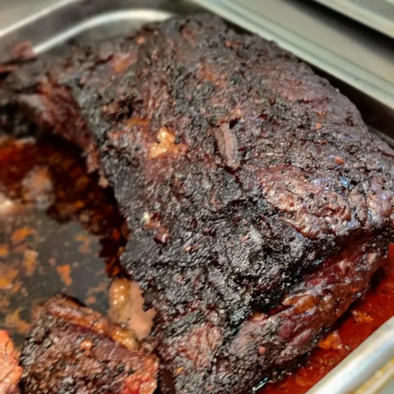 Dark, crusty smoked meat in pan with juices and sliced pieces.