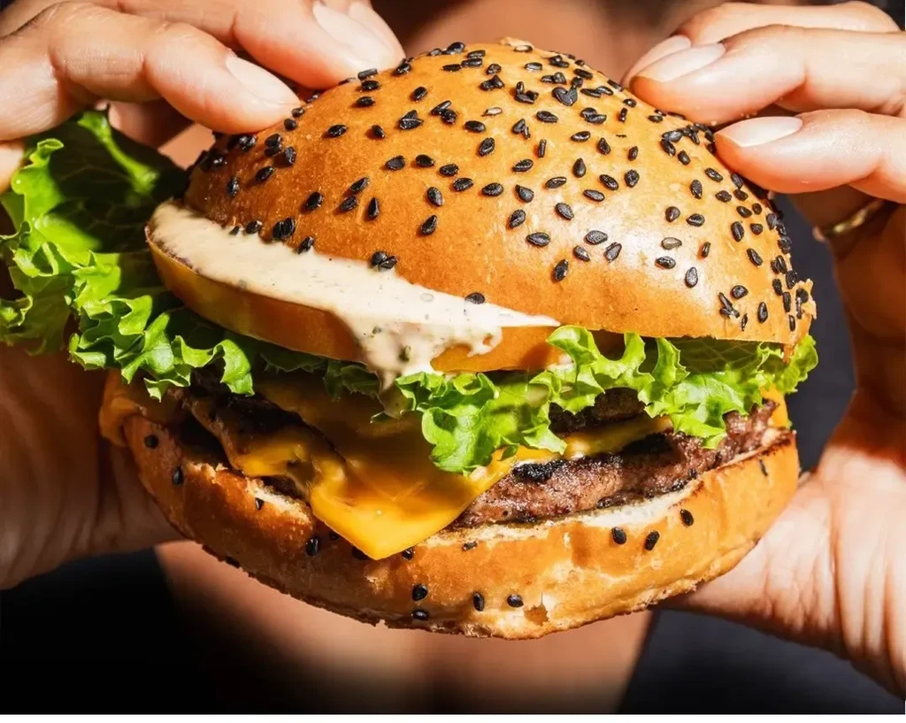 Close-up double cheeseburger with lettuce, tomato, and melted cheese, held by hands.