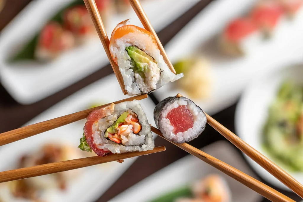 Three different sushi rolls held by chopsticks against a blurred sushi background.