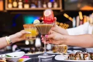 Hands toasting colorful drinks, sushi, and skewers at a bar.