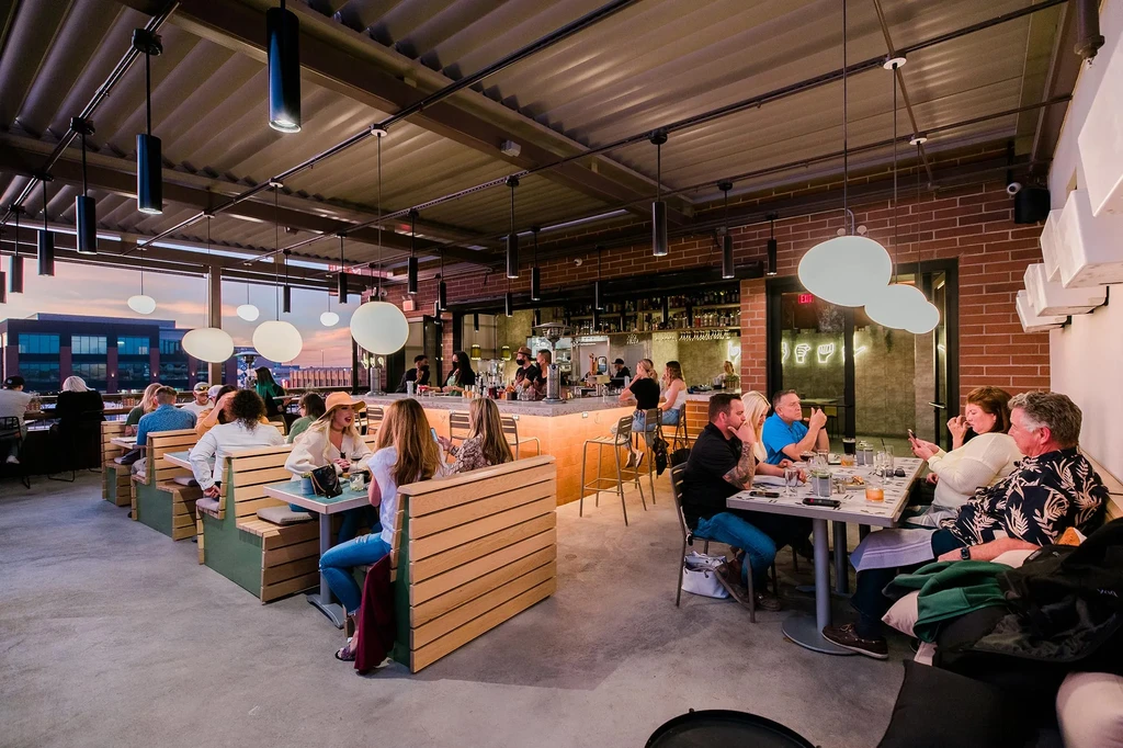 Lively industrial-chic restaurant and bar at dusk, full of patrons.