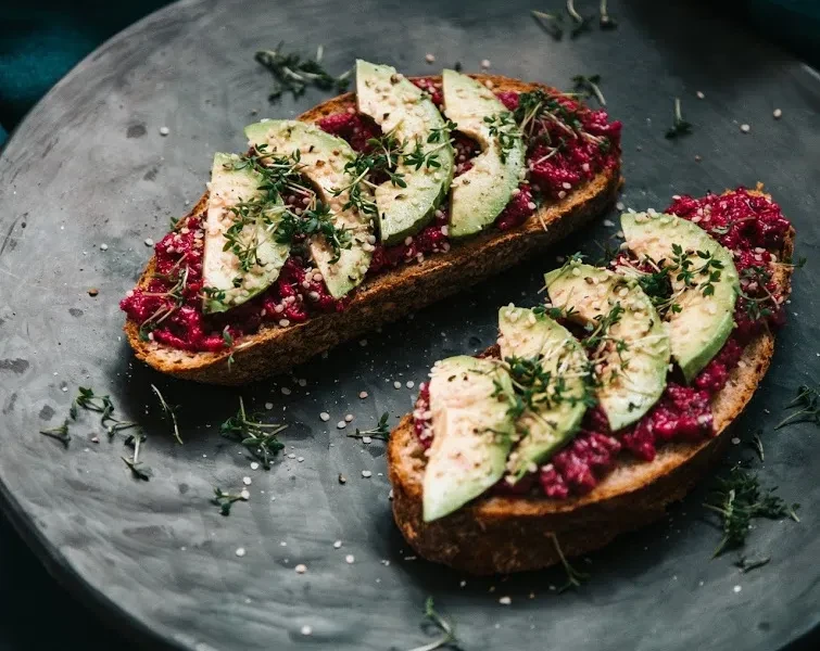 Two avocado toasts with purple spread, green avocado, herbs, and seeds.