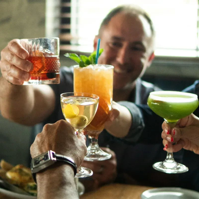 Four diverse cocktails being toasted by multiple hands in a bar.