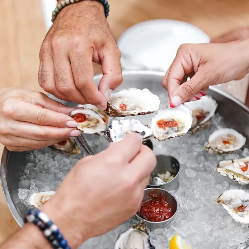 Close-up of hands reaching for fresh oysters on ice with sauces.