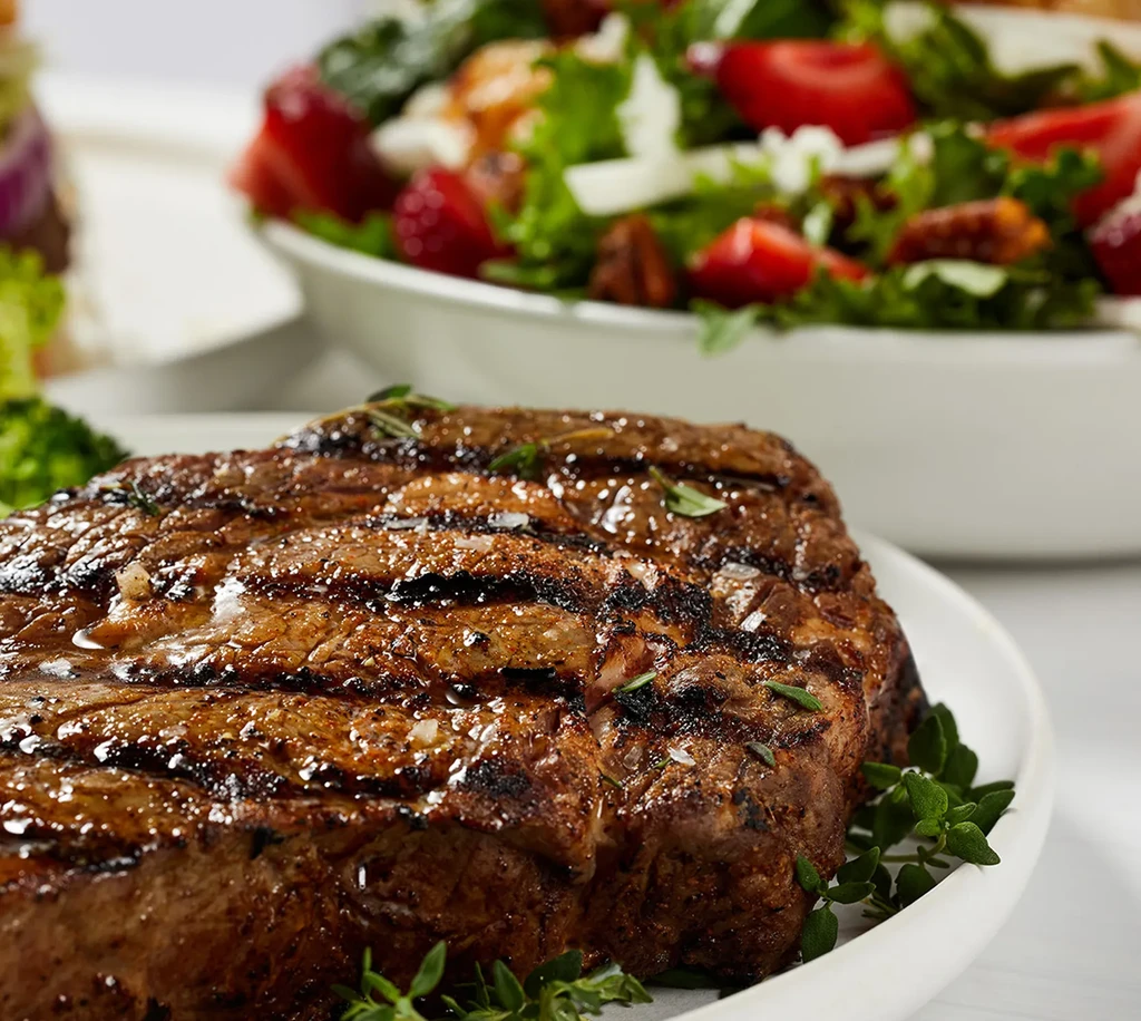 Thick grilled steak with herbs, salad bowl in soft focus background.