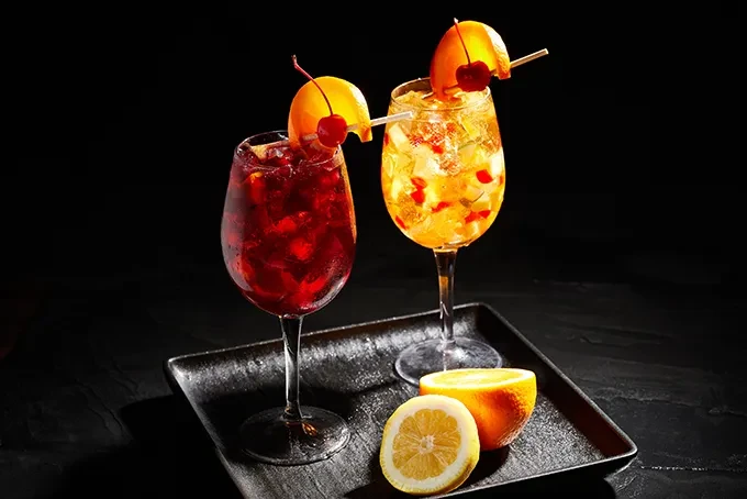 Two ruby and golden cocktails garnished with fruit on a dark tray.