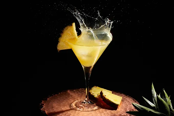 Golden cocktail in martini glass with ice, pineapple, and captured splash.