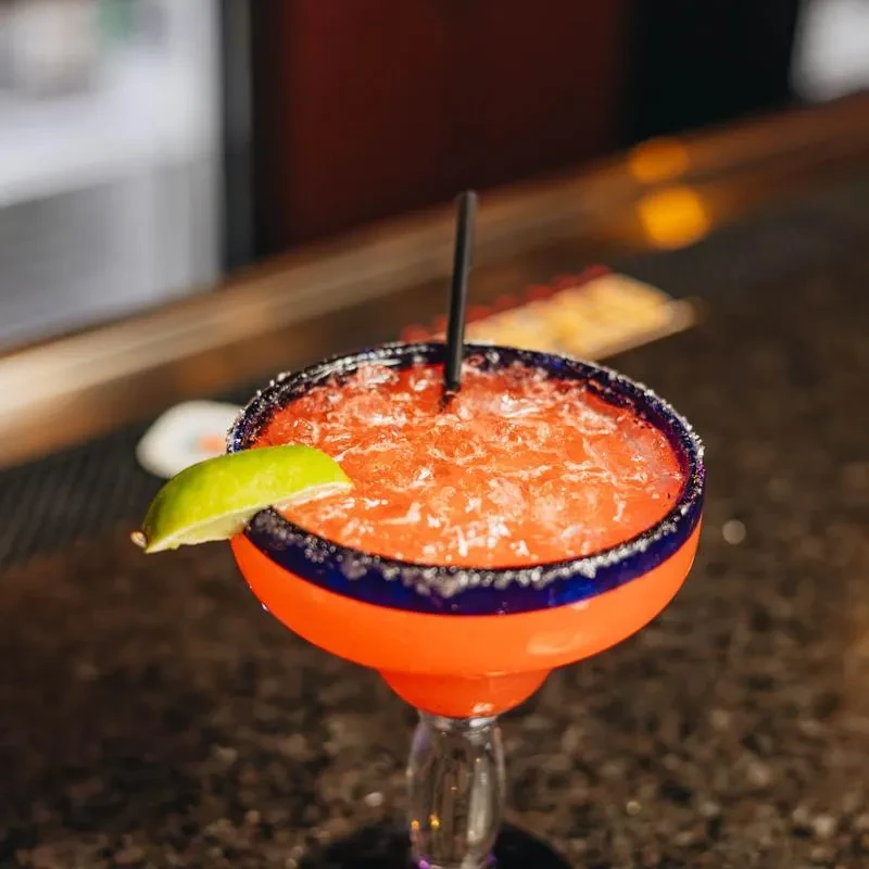 Frozen red-orange margarita with blue salted rim, lime, and straw.
