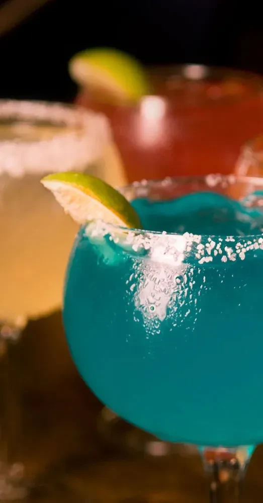 Blue cocktail with lime and salt, two other colorful drinks blurred.
