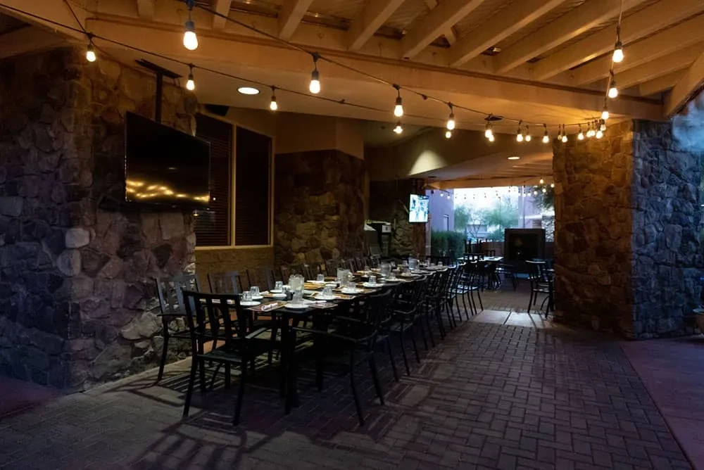Empty outdoor dining patio with long table, stone walls, string lights.