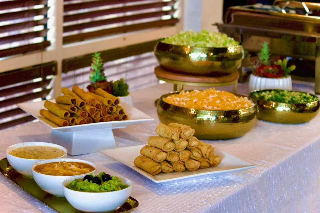 Well-arranged buffet with egg rolls, taquitos, three dips, and garnishes.