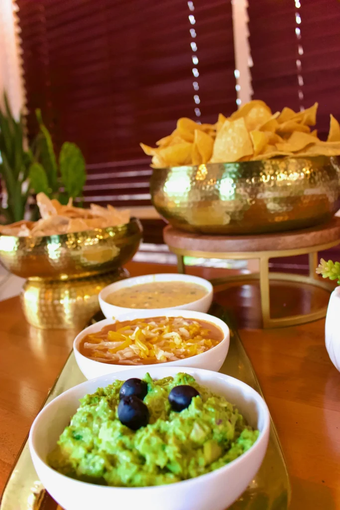Guacamole, salsa, queso, and many tortilla chips on golden serving trays.