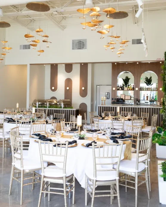 Elegant event hall with white tables, gold chairs, bar, and unique lighting.