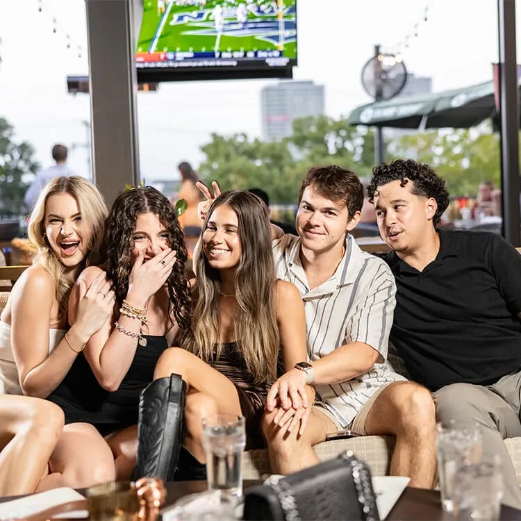 Five young adults laughing on a sports bar patio.