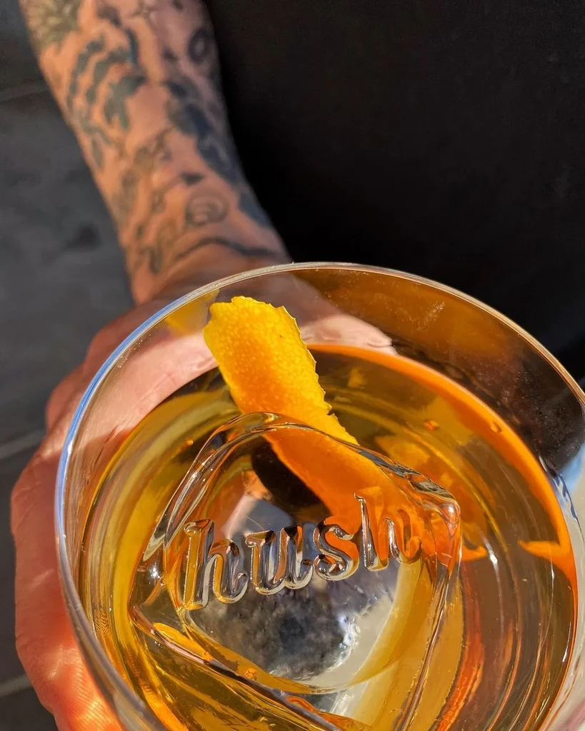 Amber cocktail with 'Hush' ice and orange peel held by tattooed hand.