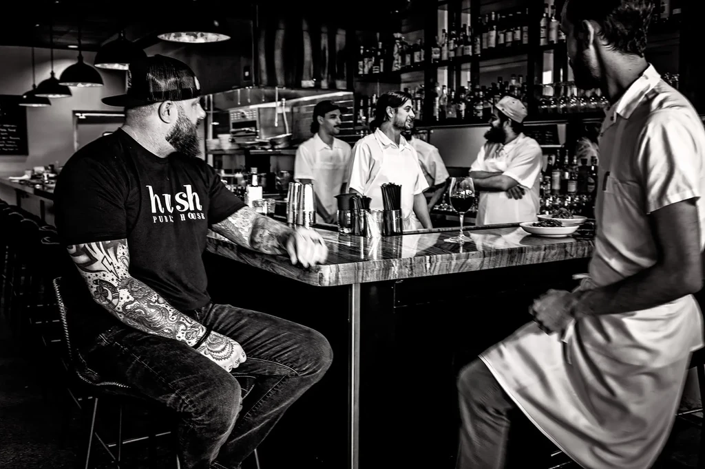 Black and white photo: patrons and staff interact at a bar.