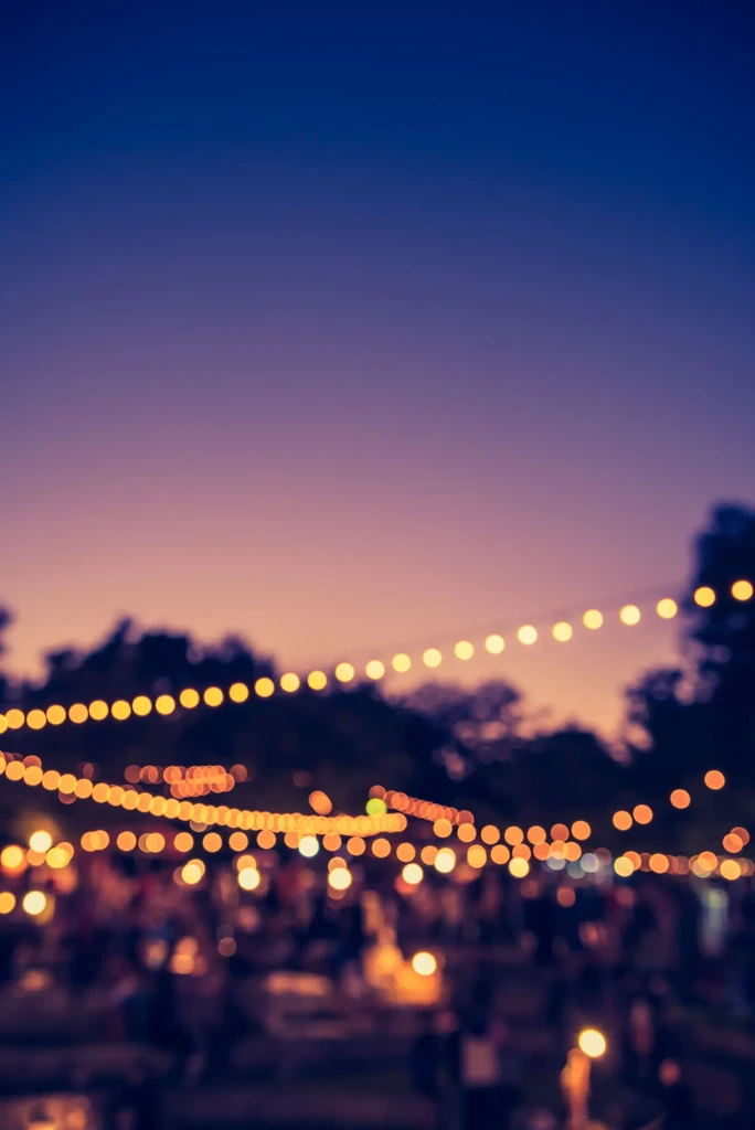 Warm bokeh string lights illuminate a colorful twilight sky above a blurred crowd.