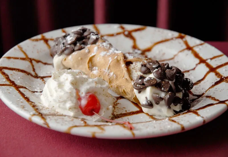 Cannoli, chocolate chips, powdered sugar, whipped cream, and cherry on white plate.