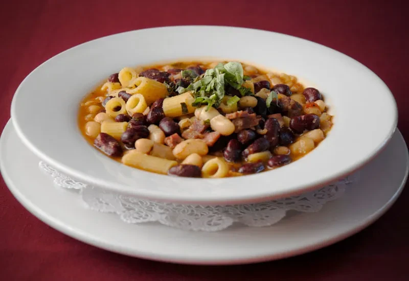 Hearty pasta and bean stew with meat and herbs in a white bowl.