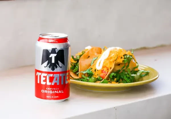 Red Tecate beer can beside three hard-shell tacos with toppings.