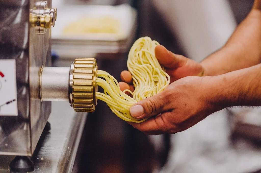 Tanned hands hold fresh spaghetti extruding from a brass-die pasta machine.