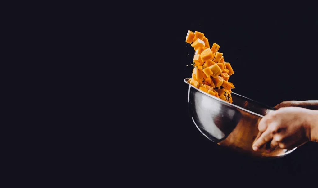 Two hands toss diced orange food with herbs from a silver bowl.