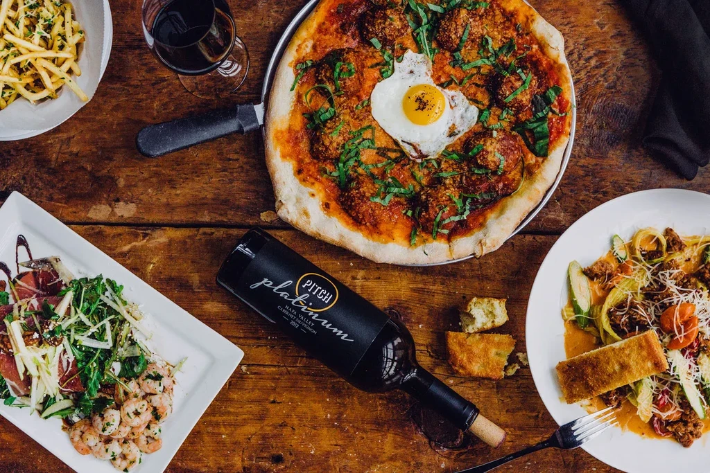 Rustic table with pizza, pasta, salad, fries, and a bottle of wine.