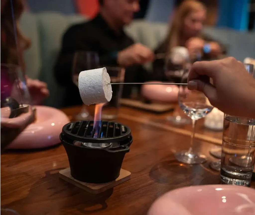 Hand roasts marshmallow over tabletop grill, restaurant background.