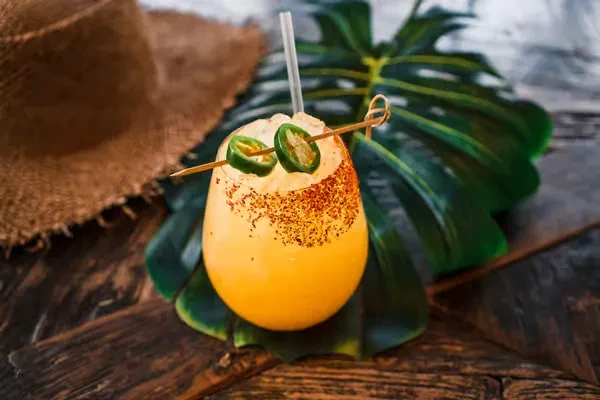 Yellowish-orange cocktail with spice rim, jalapeños, on a tropical leaf.