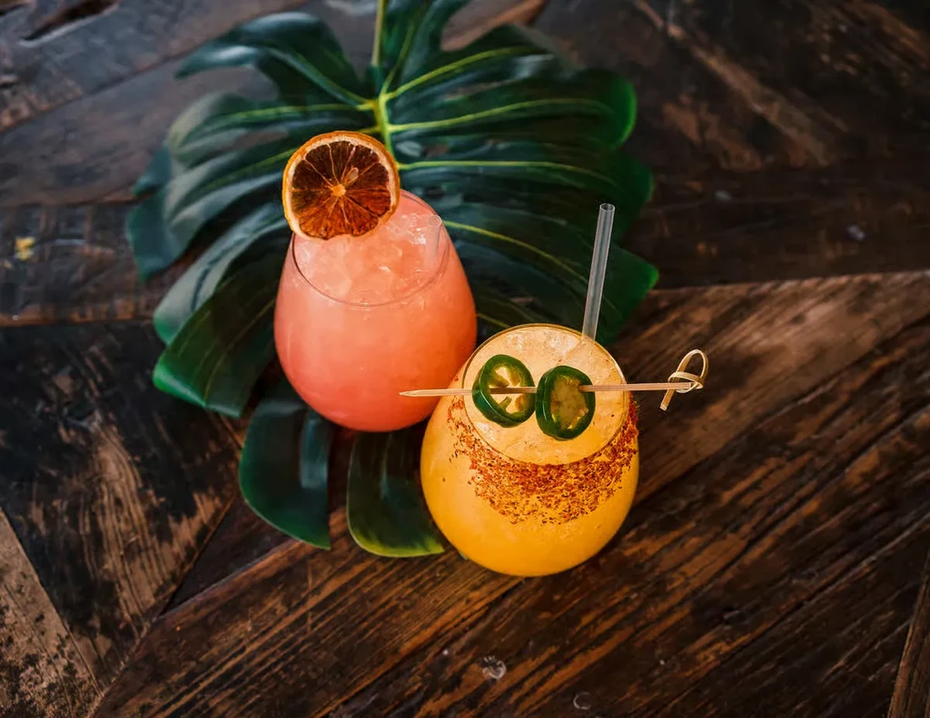 Two colorful cocktails, red and yellow, on wood with a tropical leaf.